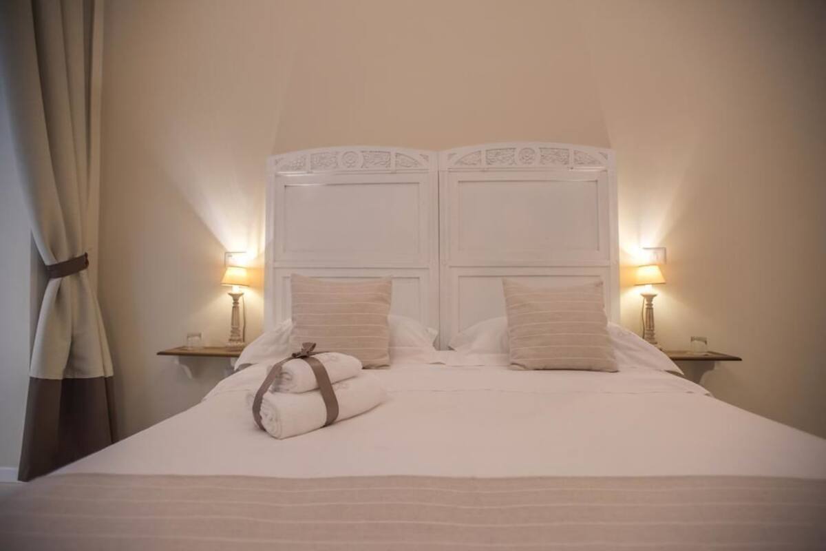 Una cama bien hecha con almohadas y una toalla enrollada, dos lámparas de noche y una cortina a la izquierda.
