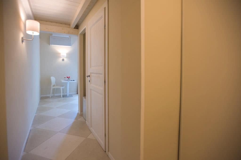 A narrow hallway with a white door on the right, a small table with a chair in the background, and a wall-mounted light fixture.