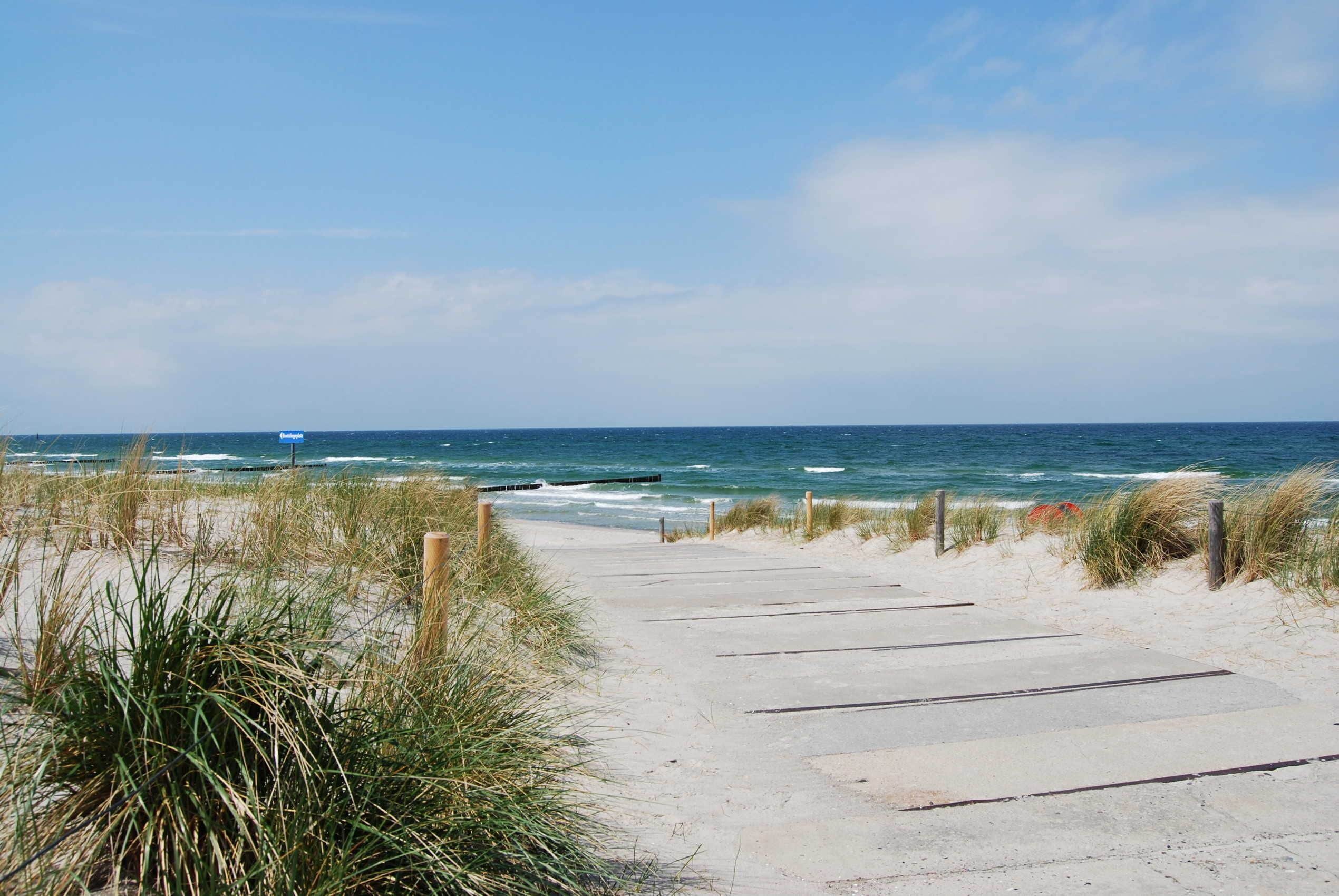 Un chemin en béton mène à la plage, avec du sable et de l’herbe de chaque côté.