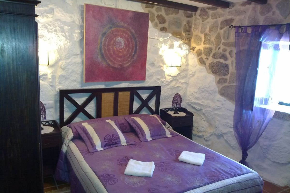Une chambre avec un mur en pierre, un lit avec une literie violette, un tableau au-dessus du lit et une table de chevet avec une lampe.