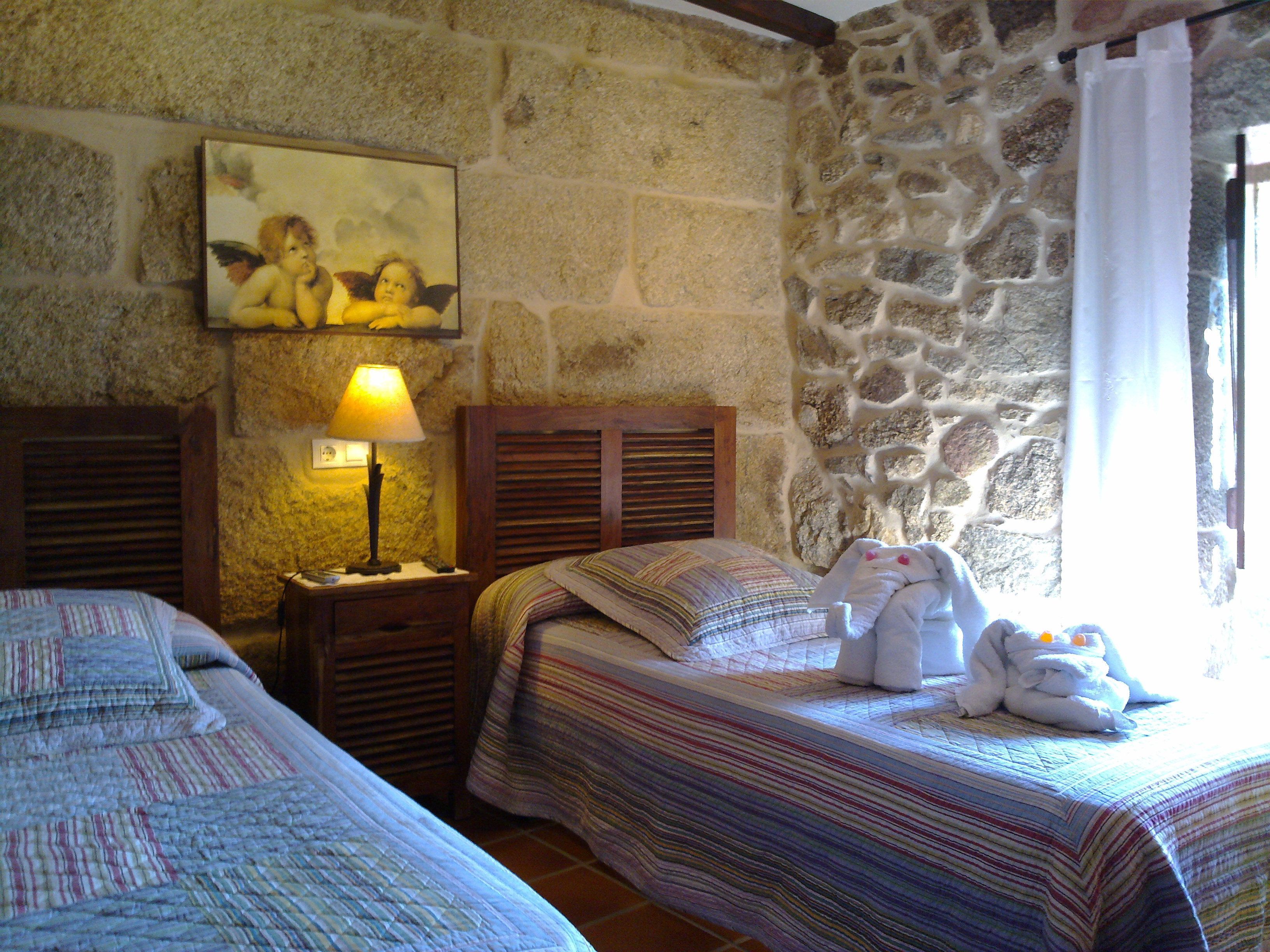 Une chambre avec deux lits, un mur en pierre, un tableau, une lampe et une fenêtre avec un rideau.