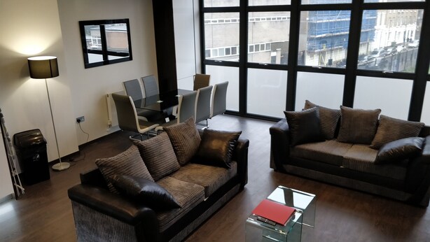A modern living room with two dark brown sofas, a glass coffee table, and a floor lamp. A large window offers a view of the cityscape. - Access Tower Hill (London)