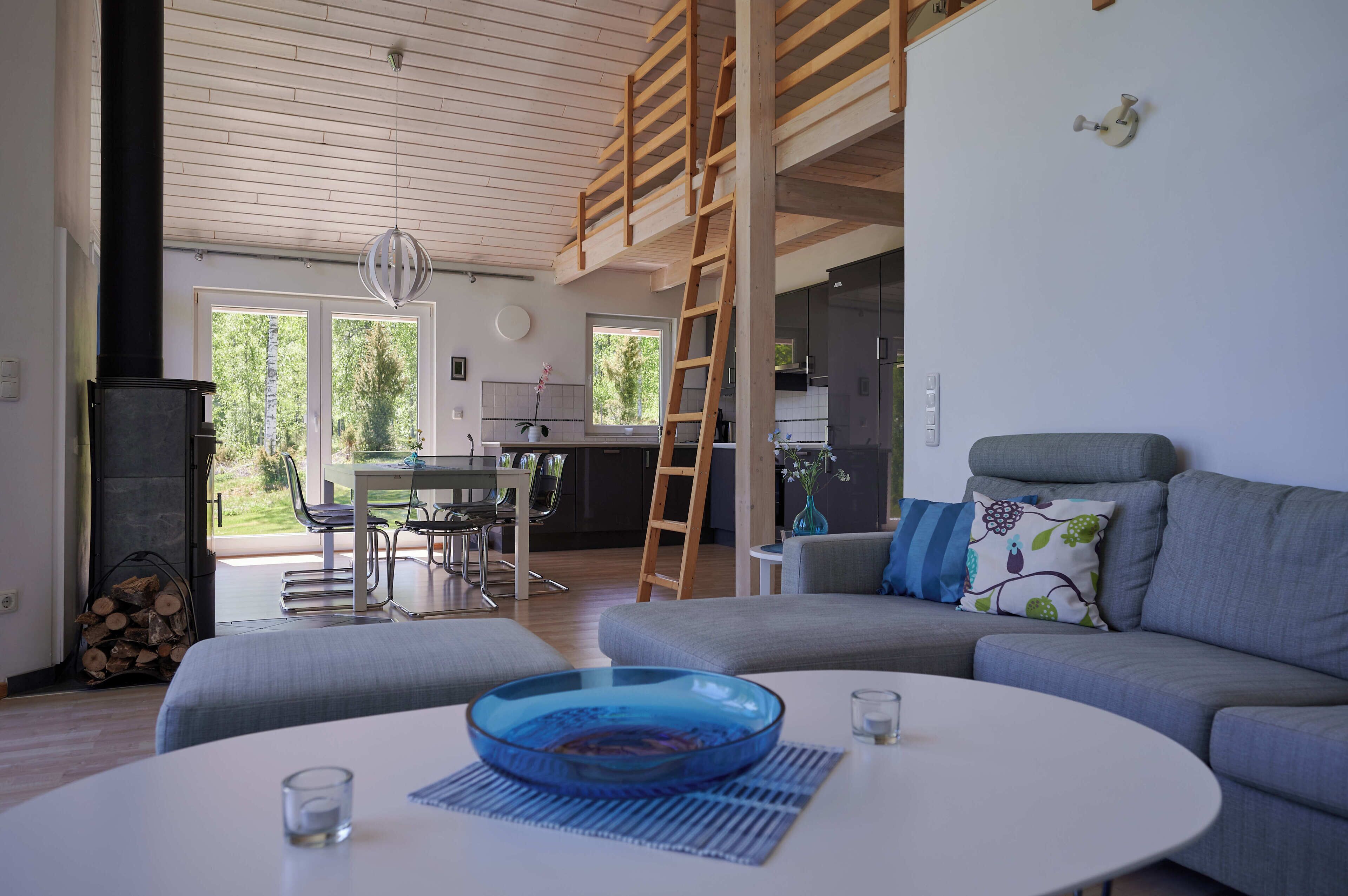 Una sala de estar moderna con un sofá gris, una mesa de centro redonda con un cuenco de vidrio azul, y una zona de comedor con una mesa y sillas de vidrio.