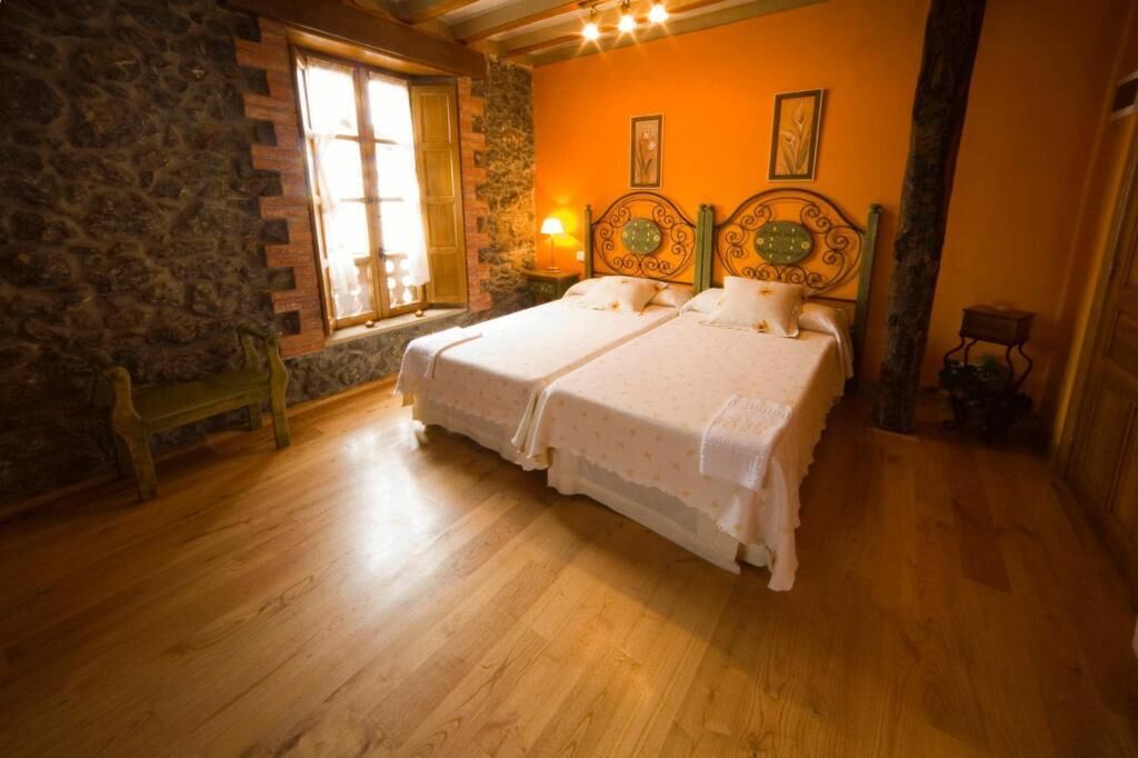 A room with a stone wall, wooden floor, two beds with white bedding, a bench, a window with a view, and a wall-mounted lamp.
