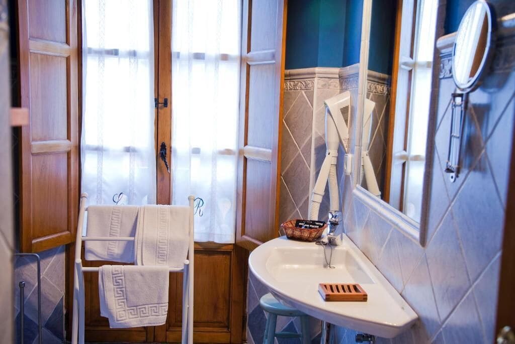 Un baño con un lavamanos blanco, un espejo, un perchero con toallas y una ventana con cortina.