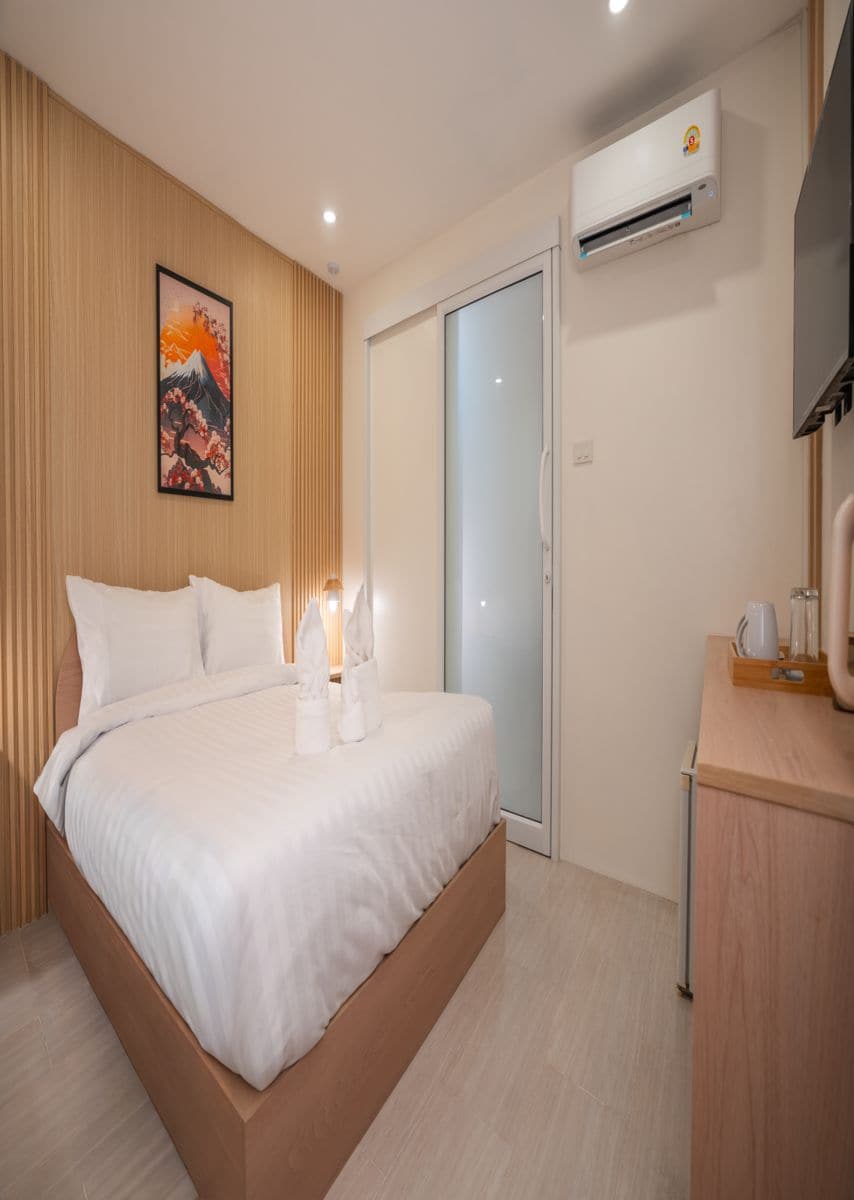 Habitación de hotel moderna con cama, cabecera de madera, aire acondicionado de pared, puerta corredera de vidrio y mesita de noche con lámpara.