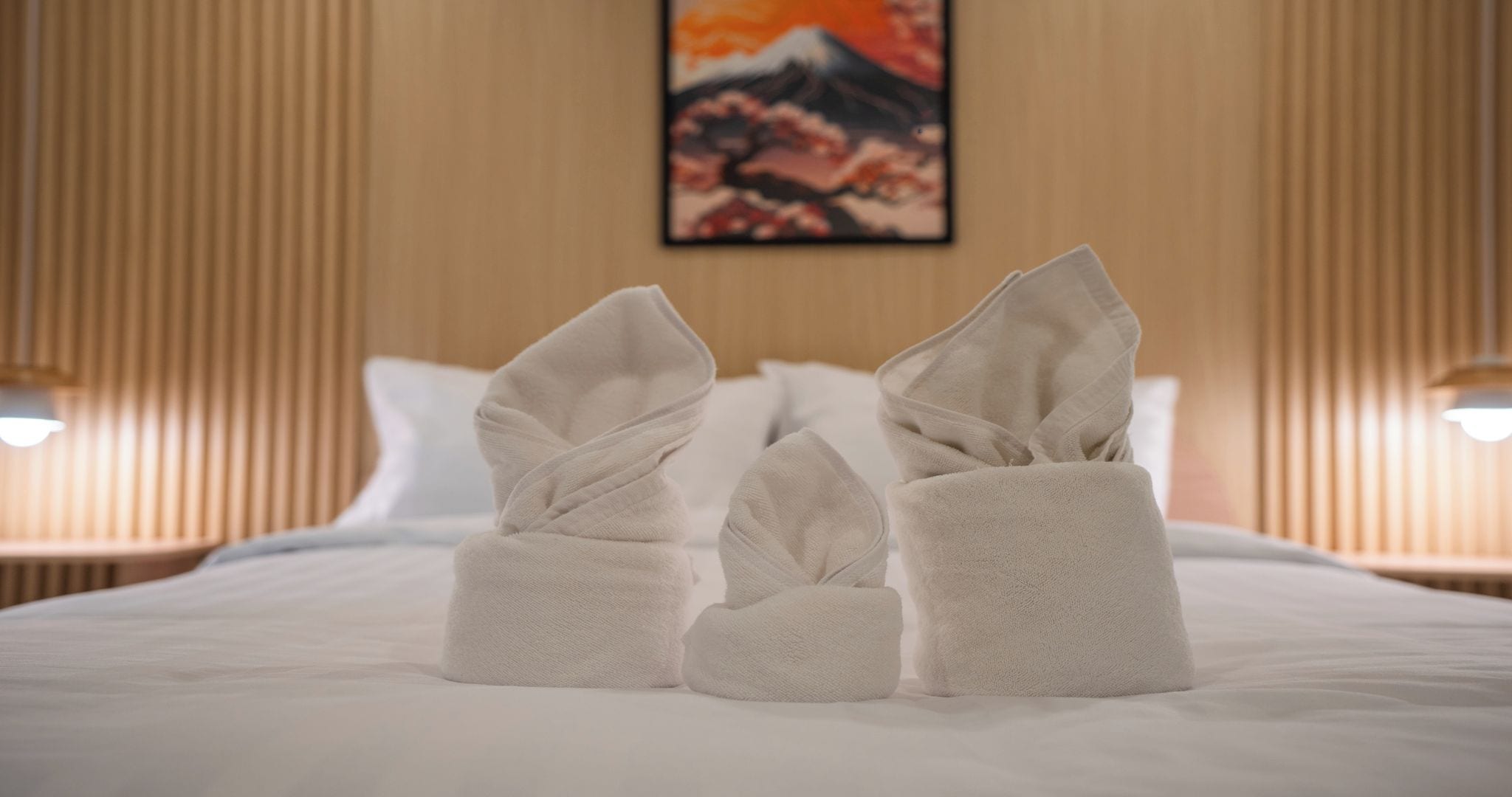 Una cama bien hecha con toallas dispuestas de forma decorativa, un cuadro en la pared y dos lámparas de noche.