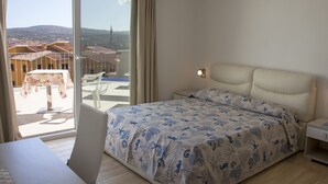 A bedroom with a bed, a desk, and a view of the city through large windows. - Rena Bianca Suite (Santa Teresa di Gallura)
