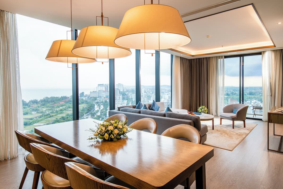 A modern hotel room with a dining area, large windows with a city view, and a sofa set.