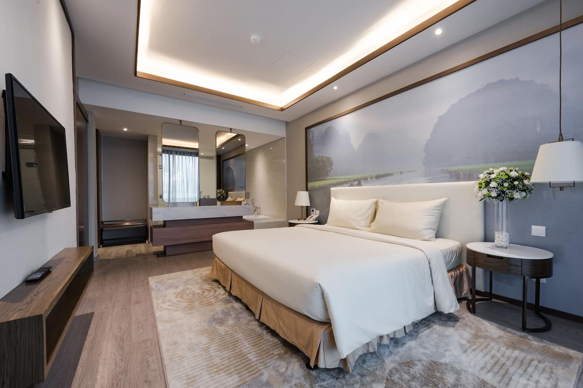 A modern hotel room with a large bed, a TV, a bathroom with a mirror, and a painting on the wall.