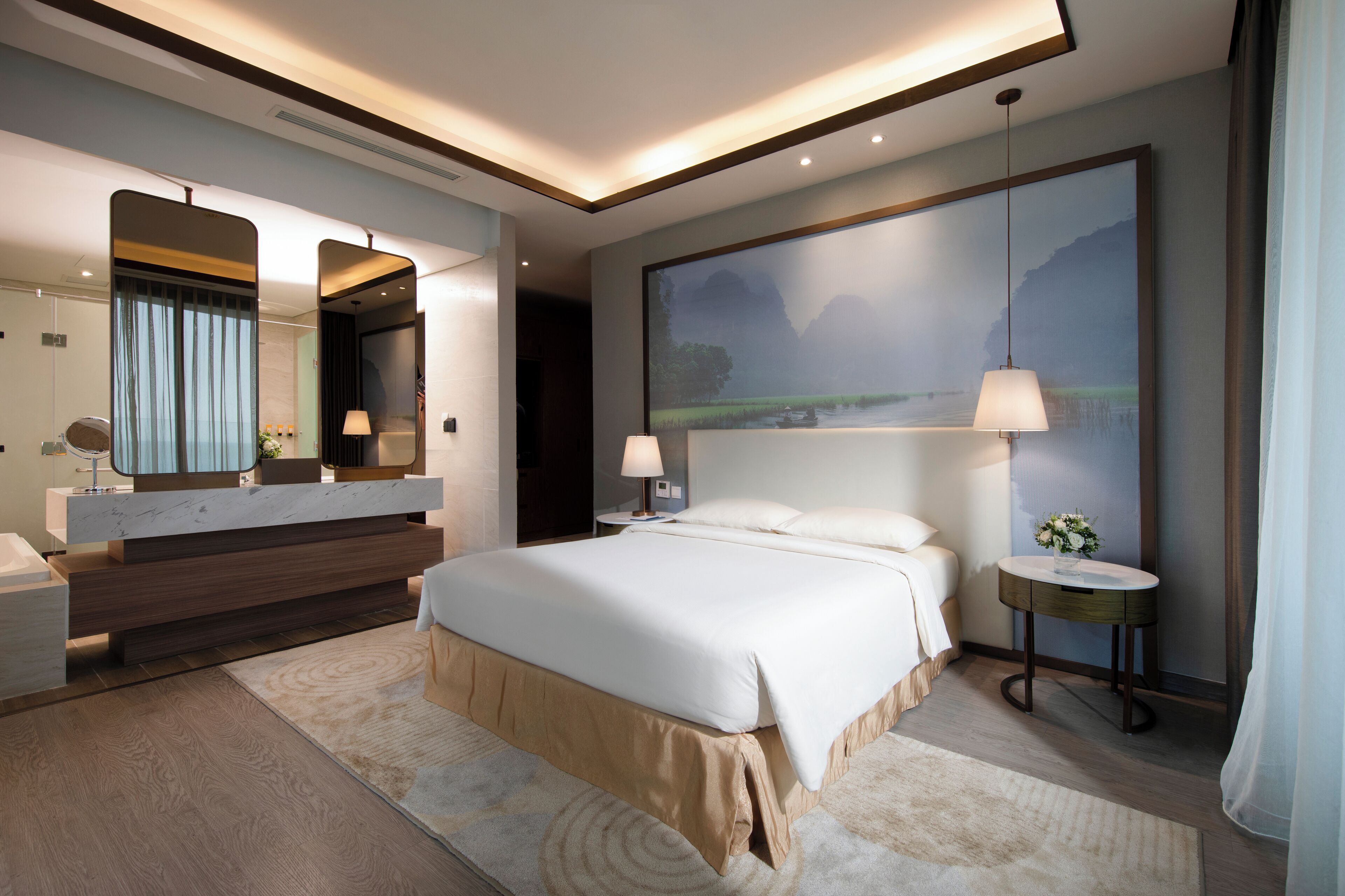 Een moderne hotelkamer met een groot bed, een kaptafel met spiegel, een rond bijzettafeltje en een groot schilderij van een landschap.