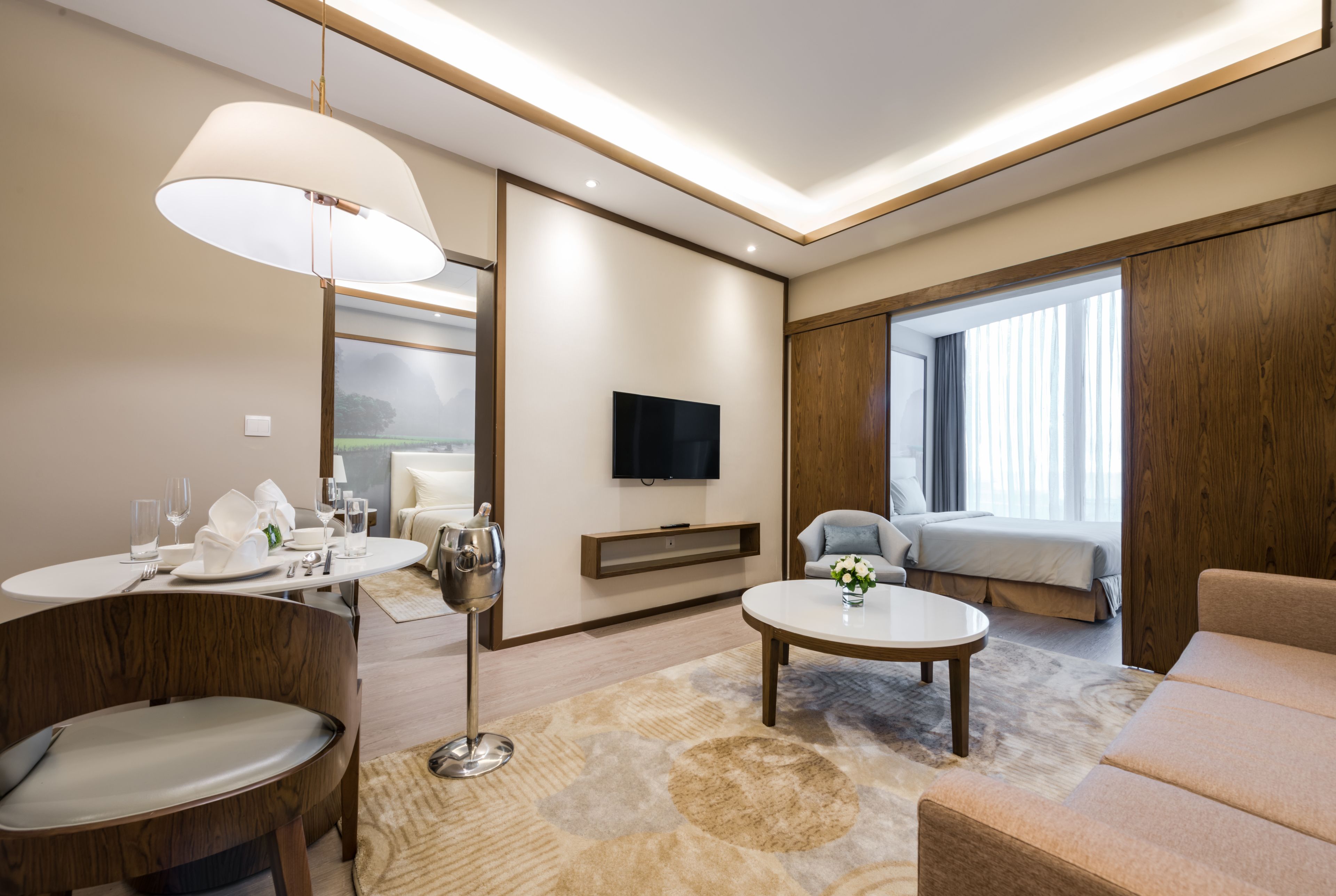 A modern hotel room with a dining area, a sofa, a TV, and a large window.