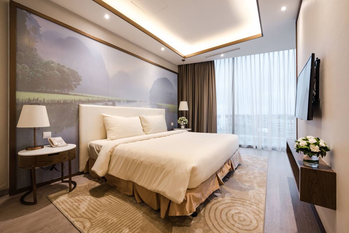 A modern hotel room with a large bed, a bedside table with a lamp, a small table with a phone, a large window with curtains, and a wall-mounted TV.