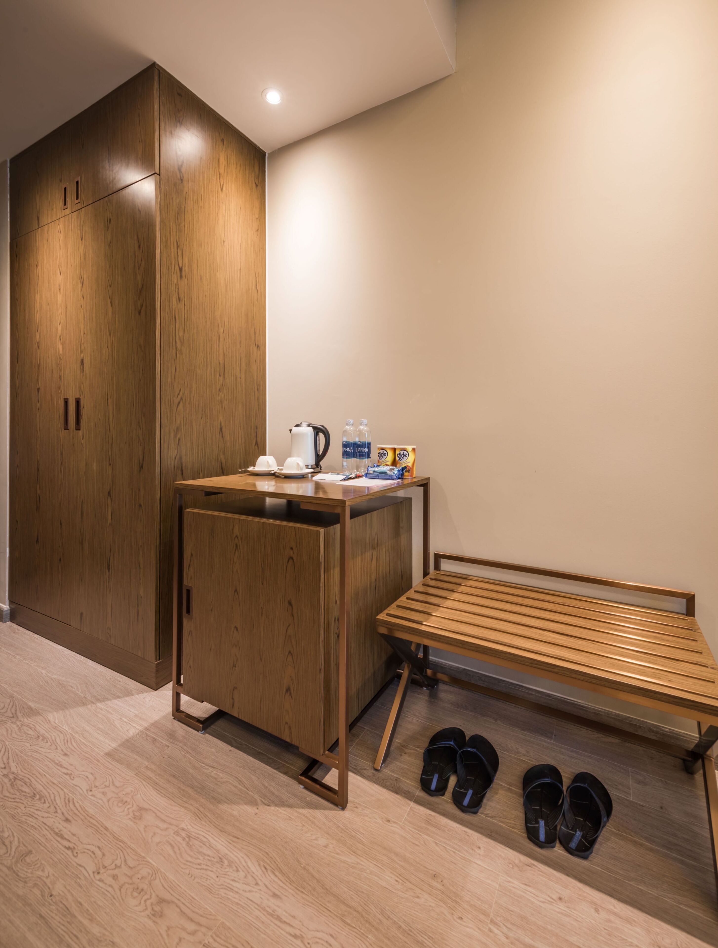 A room with a wooden wardrobe, a small table with a kettle, cups, and snacks, a bench, and a pair of slippers on the floor.