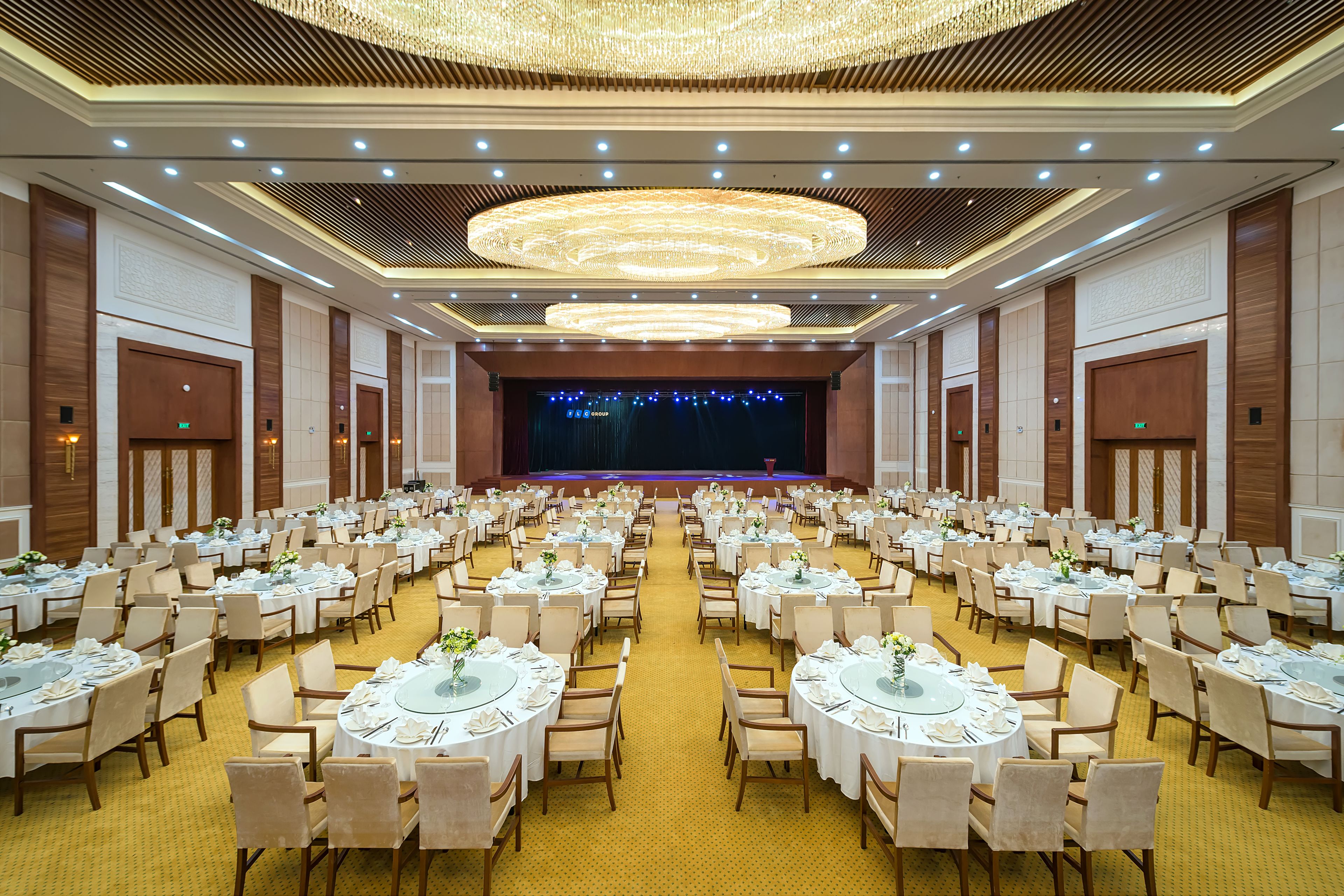 A large banquet hall with round tables, chairs, and a stage at the front.