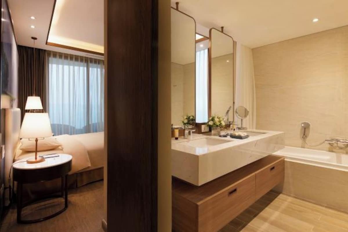 A modern hotel bathroom with a large mirror, two sinks, a bathtub, and a bedside table with a lamp.