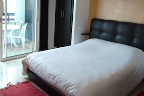 A bedroom with a large bed, a sliding glass door leading to a balcony with chairs, and a dark-colored wardrobe. - Marina Agadir Maliari (Agadir)