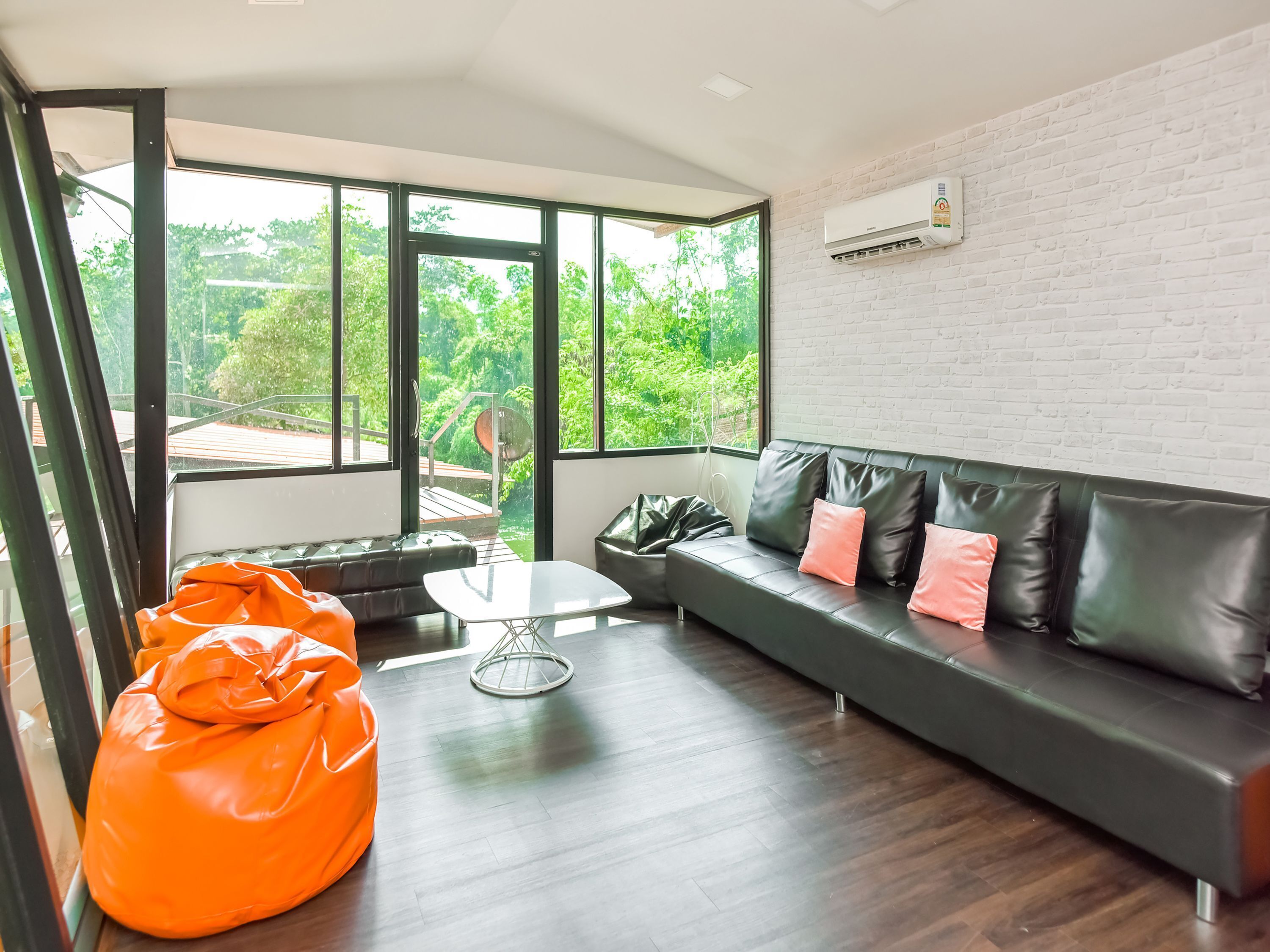 A modern living room with a black leather sofa, a glass coffee table, and an orange bean bag. Large windows offer a view of greenery outside.
