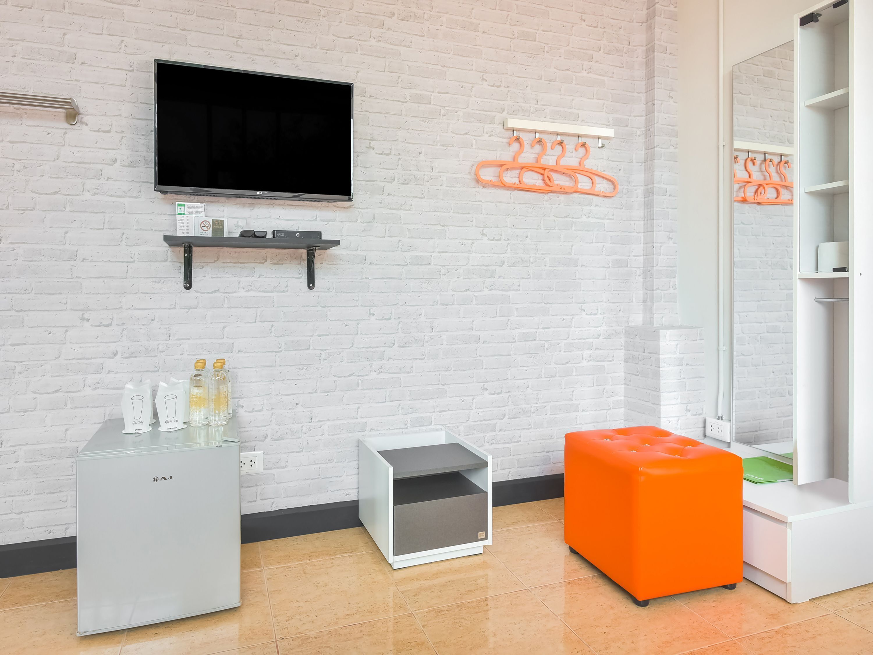 Une chambre moderne avec un mur en briques blanches, un téléviseur à écran plat, un mini-réfrigérateur, un petit meuble et un pouf orange.