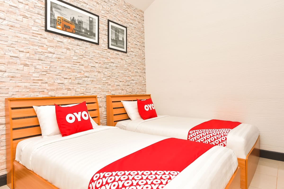 Une chambre d’hôtel avec deux lits, chacun doté d’une tête de lit en bois, de draps blancs et d’oreillers rouges ornés du logo « OYO ». La chambre possède un mur avec un motif en briques et un tableau encadré au-dessus des lits.