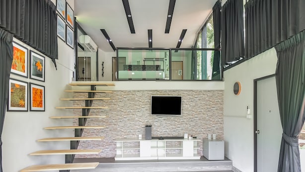 A modern interior with a staircase leading to a balcony, a flat-screen TV mounted on a stone wall, and a minimalist reception desk. - Nana River Kaeng Krachan (Kaeng Krachan)