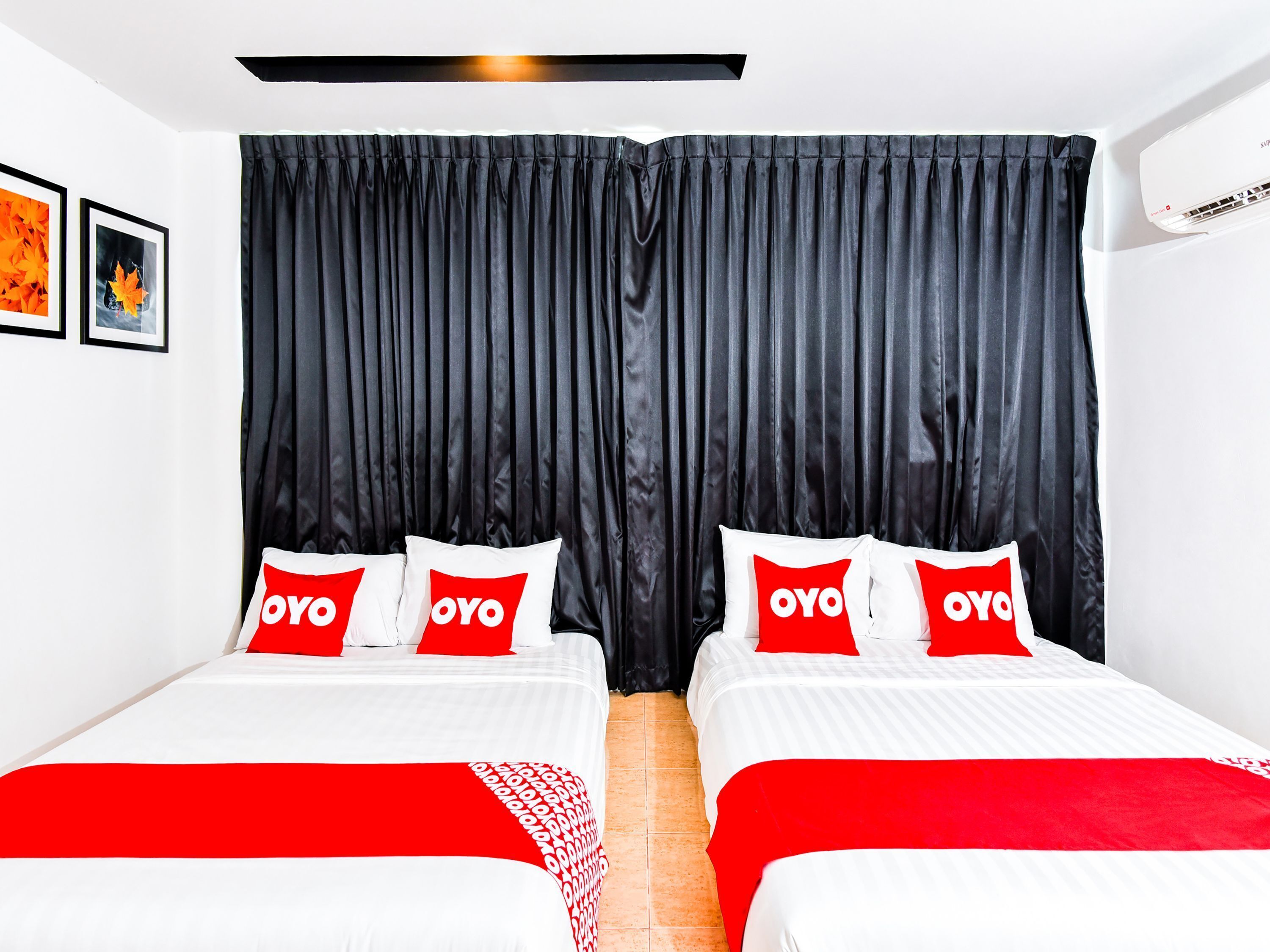 A hotel room with two beds, each with red and white bedding and pillows featuring the 'OYO' logo. The room has a large black curtain, white walls, and a tiled floor.