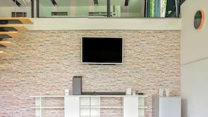 A modern living room with a stone wall, a flat-screen TV mounted above a white entertainment unit, and a small white speaker. - Nana River Kaeng Krachan (Kaeng Krachan)