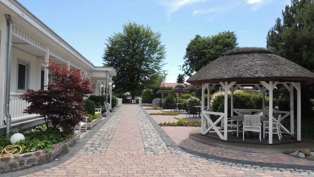 A paved pathway leading to a gazebo with outdoor seating, surrounded by greenery and a building with a white facade. - Sunshine (7) - Landhaus Mönkebude (Mönkebude)
