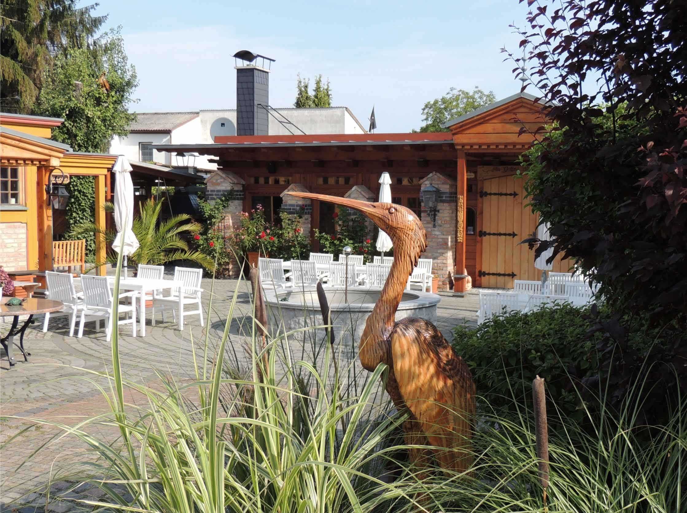En udendørs terrasse med hvide stole og borde, et træpergola og en stor fugleskulptur.