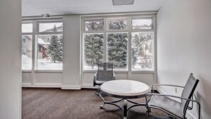 A conference room with a round table and two chairs, large windows with a view of snow-covered trees and a building. - Vs440h Village Square Hotel Room (Copper Mountain)