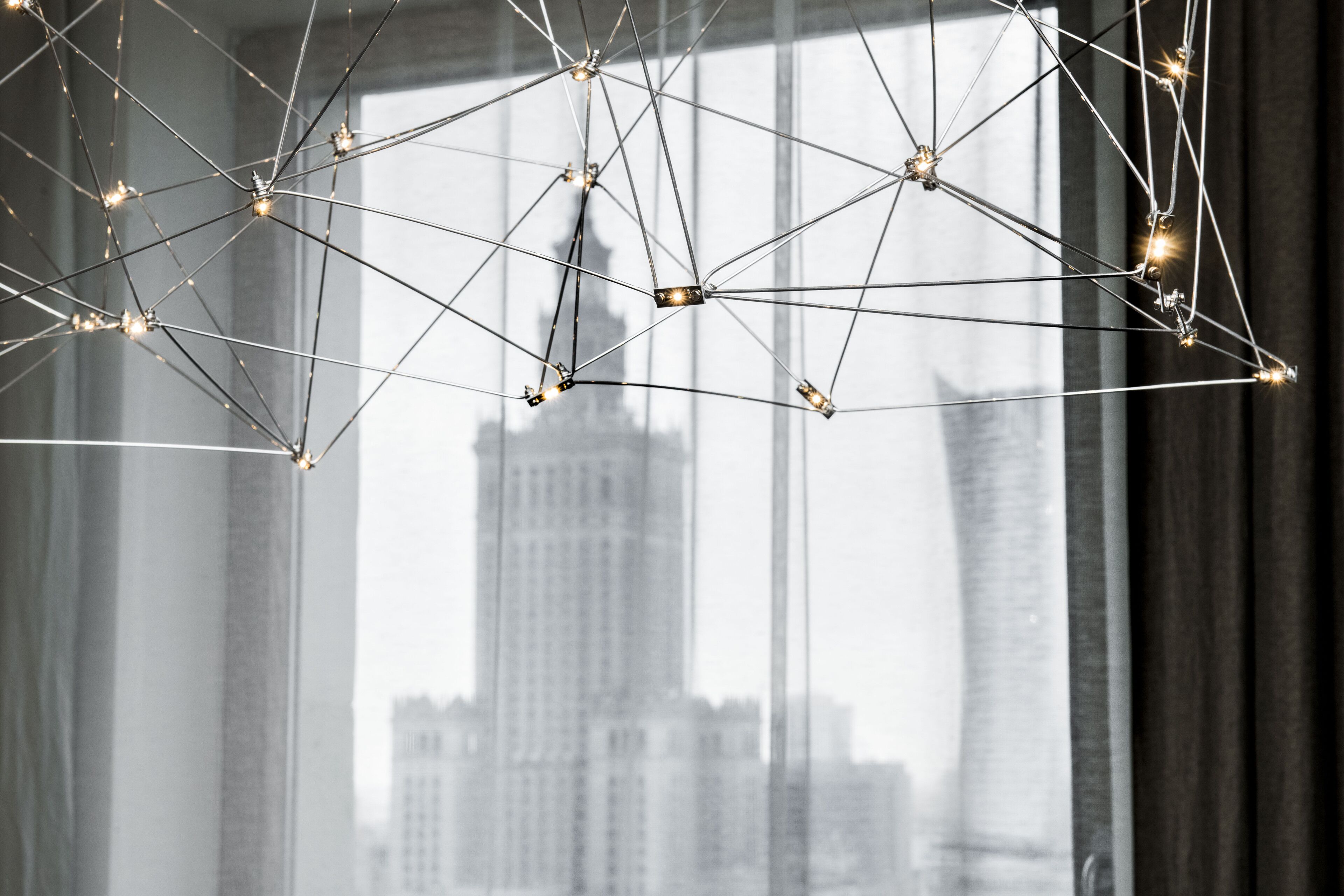 A modern chandelier with a geometric design, hanging in front of a window with a cityscape view.