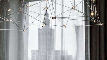 A modern chandelier with a geometric design, hanging in front of a window with a cityscape view.