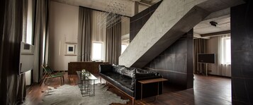 Un salon moderne avec un canapé en cuir noir, une table basse en verre et un escalier adossé à un mur en béton.