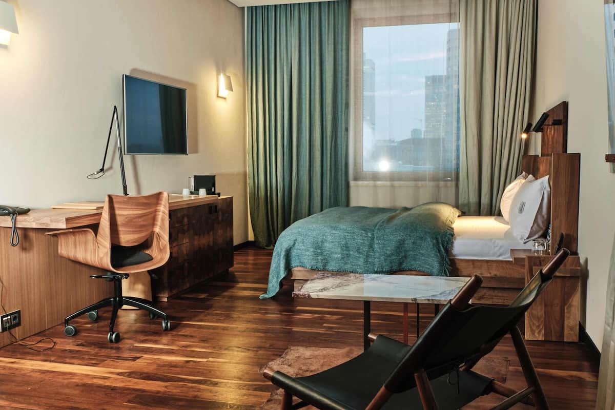 A modern hotel room with a wooden floor, a desk with a chair, a bed with a green blanket, and a large window with curtains.