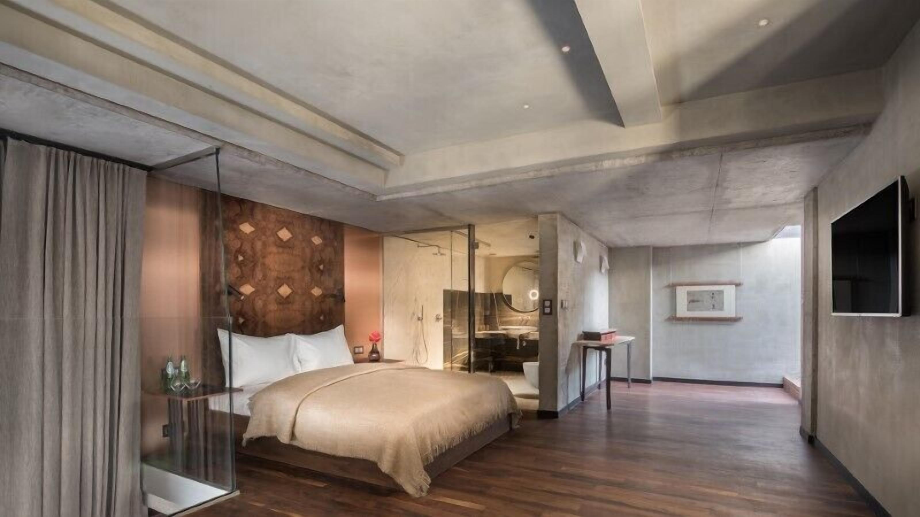 A modern hotel room with a large bed, a glass partition, a wooden headboard, a shower area, and a mounted TV.