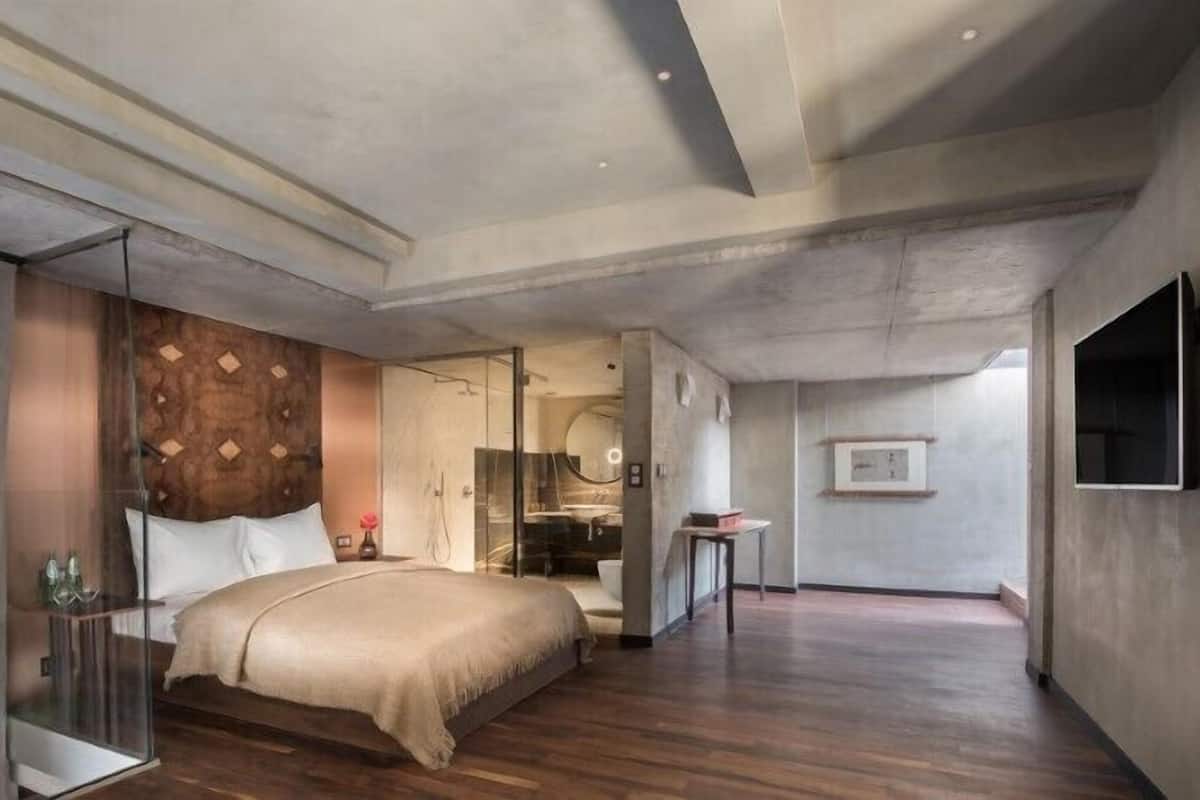 A modern hotel room with a large bed, a glass partition, a wooden headboard, a shower area, and a mounted TV.