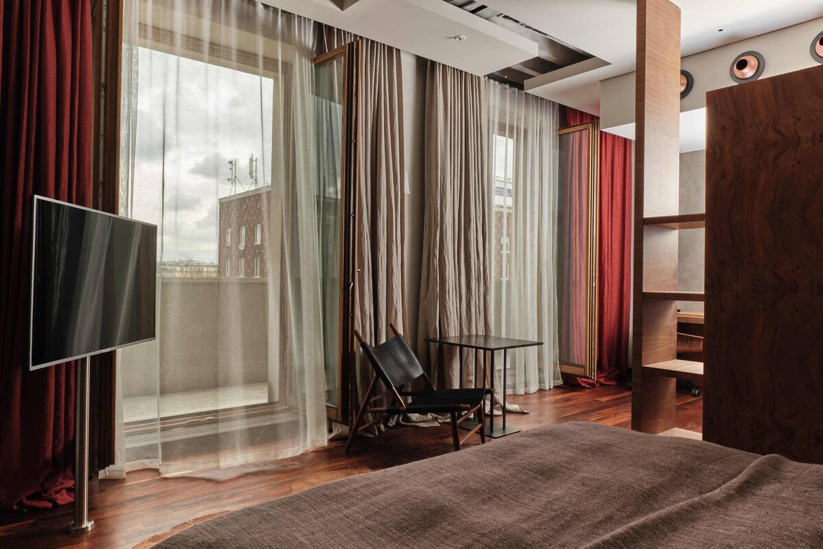 A modern hotel room with a large bed, a TV, a chair, a small table, and a view of the city through sheer curtains.