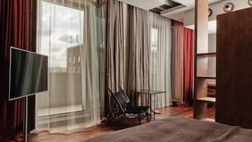Moderni hotellihuone, jossa on suuri sänky, televisio, tuoli, pieni pöytä ja näkymä kaupunkiin ohuiden verhojen läpi.