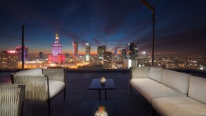 A rooftop terrace with a city skyline view, featuring a high-rise building with a distinctive spire, a modern sofa, and two small tables. - Hotel Warszawa (Warsaw)