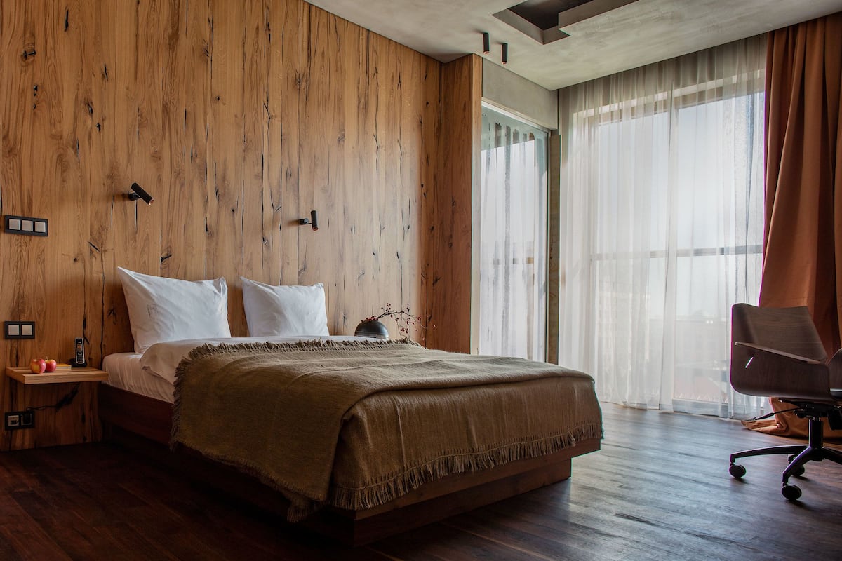 A modern bedroom with a wooden wall, a bed with white linens, a bedside table with a lamp, and a chair.