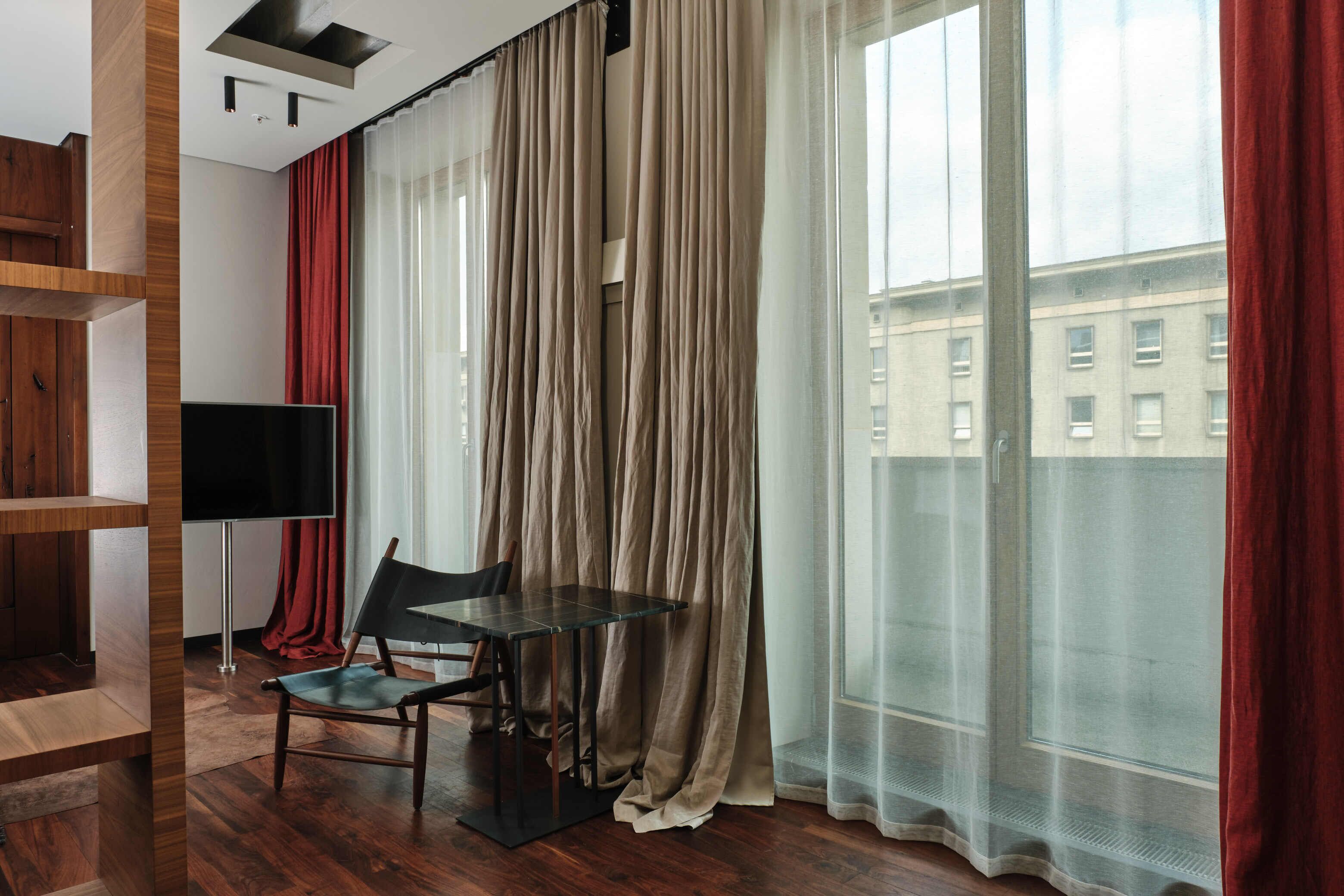 Una habitación moderna con suelo de madera, una mesa de vidrio, una silla, un televisor y ventanas grandes con cortinas translúcidas.