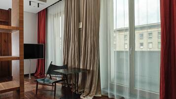 Una habitación moderna con suelo de madera, una mesa de vidrio, una silla, un televisor y ventanas grandes con cortinas translúcidas.