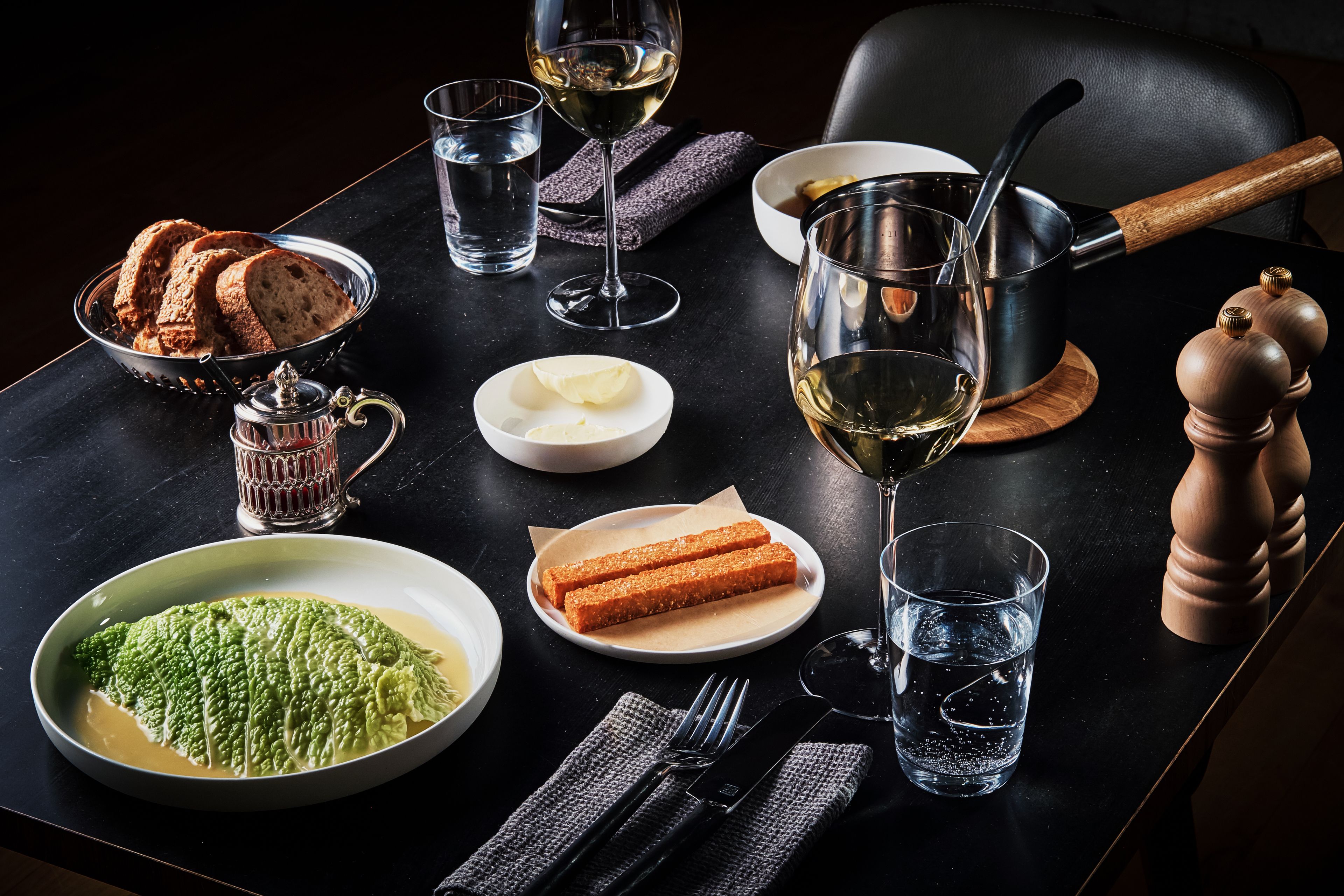 Une table à manger dressée avec du pain, du beurre, un bol de sauce verte, des verres à vin et une poêle.