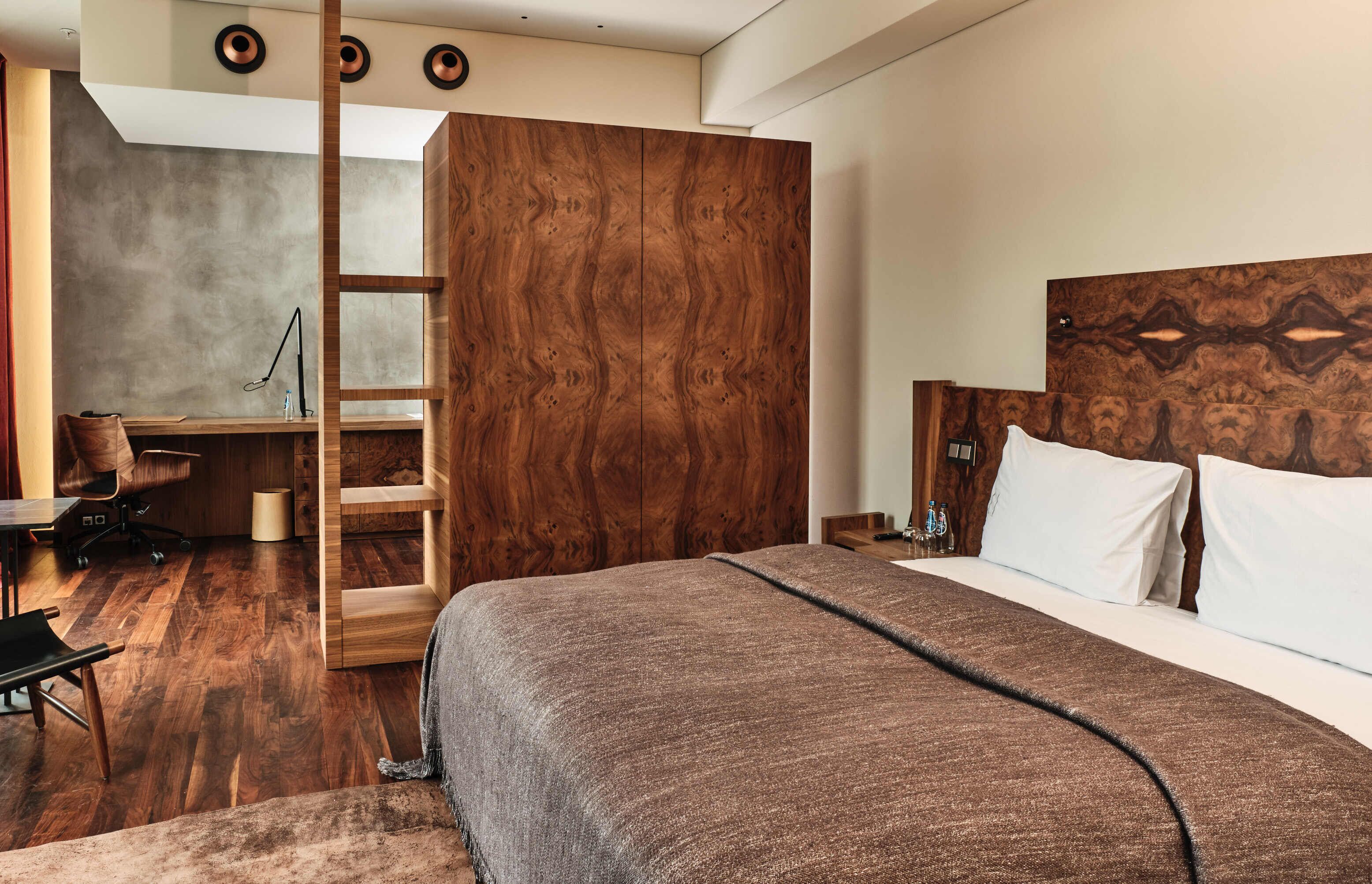 Una habitación de hotel moderna con un armario de madera, una cama con una colcha marrón, un escritorio con una silla y una mesita de noche con una lámpara.