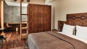 Una habitación de hotel moderna con un armario de madera, una cama con una colcha marrón, un escritorio con una silla y una mesita de noche con una lámpara.
