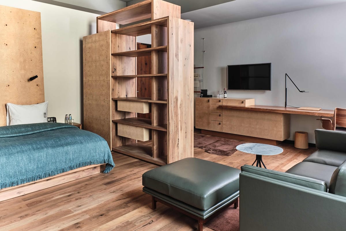 A modern hotel room with a wooden bed, a green sofa, a small round table, a wooden wardrobe, and a TV mounted on the wall.