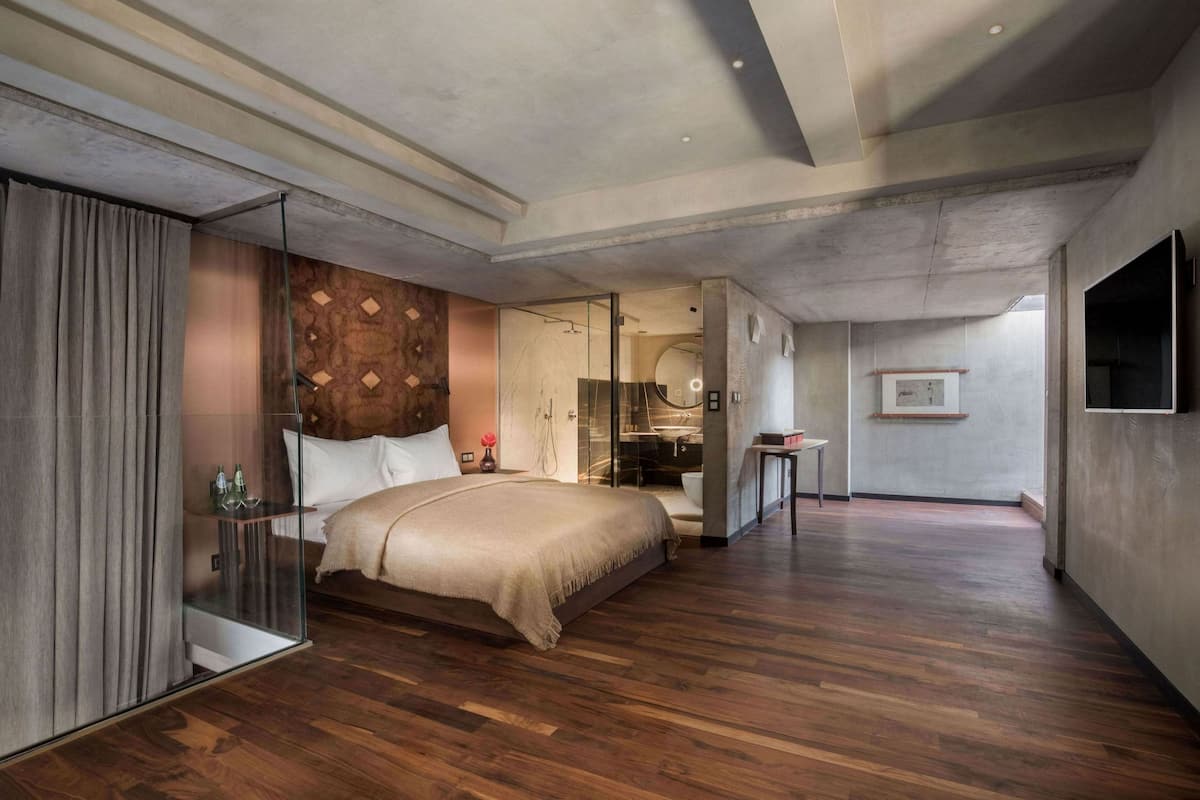 A modern bedroom with a large bed, a glass partition, a bathroom with a shower, and a TV mounted on the wall.