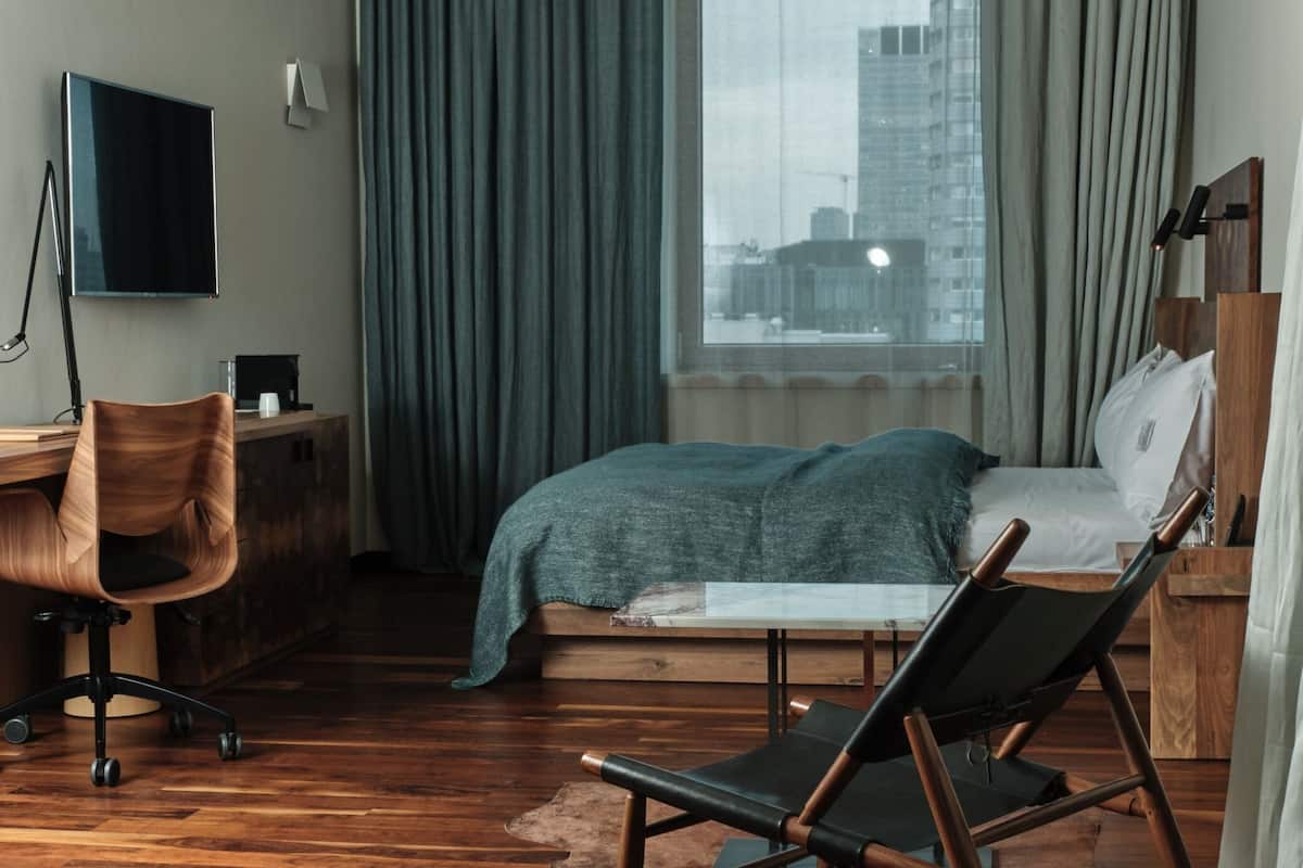 A modern hotel room with a wooden floor, a bed with a blue blanket, a desk with a chair, a TV, and a large window with curtains.