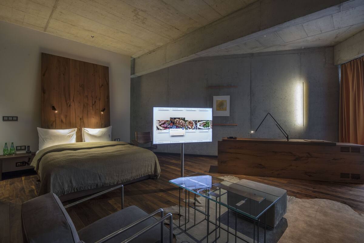 A modern hotel room with a wooden headboard bed, a glass coffee table, a desk with a computer, and a large flat-screen TV mounted on the wall.