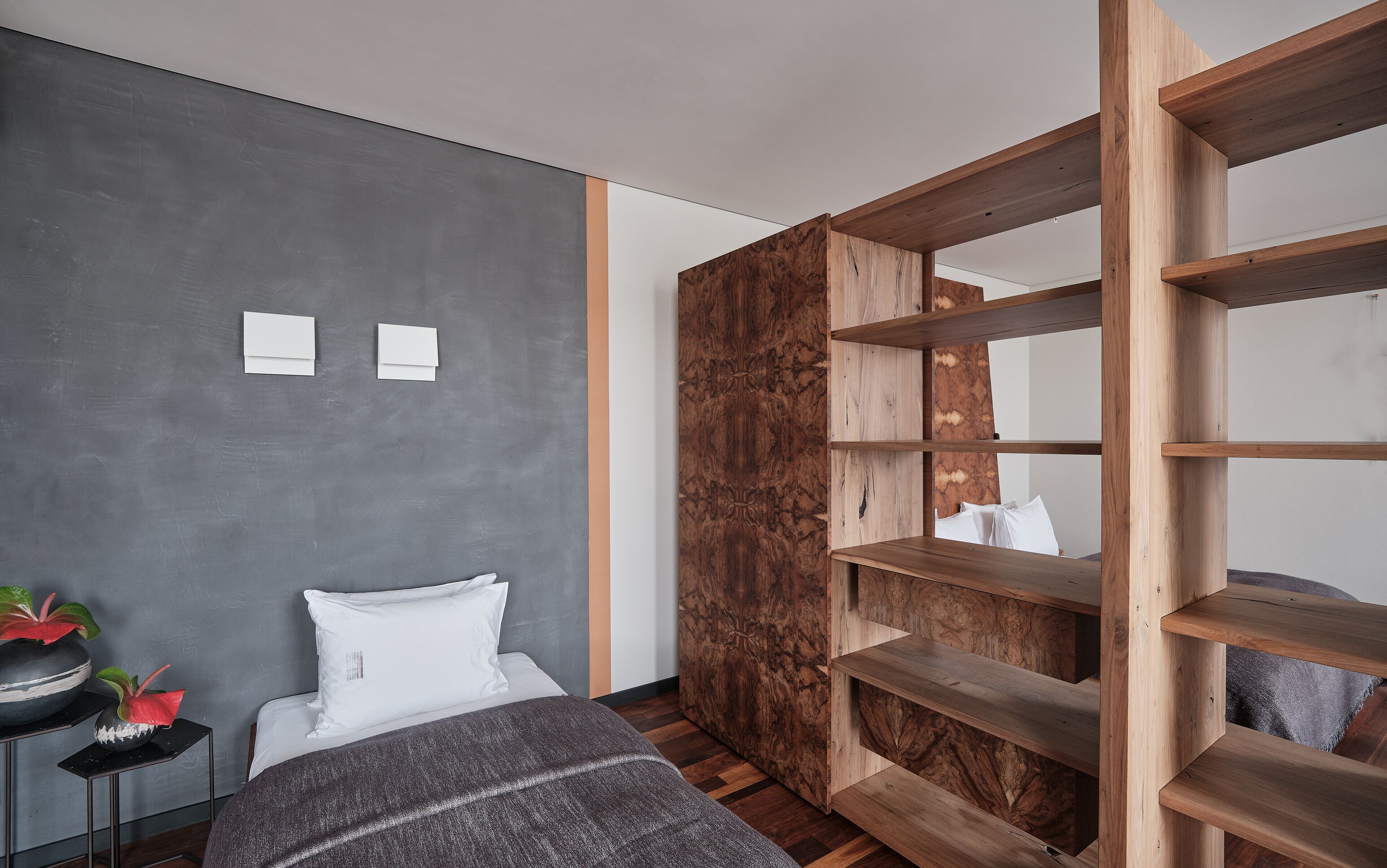 A modern bedroom with a grey wall, a wooden bookshelf, a bed with white bedding, and a small table with decorative items.